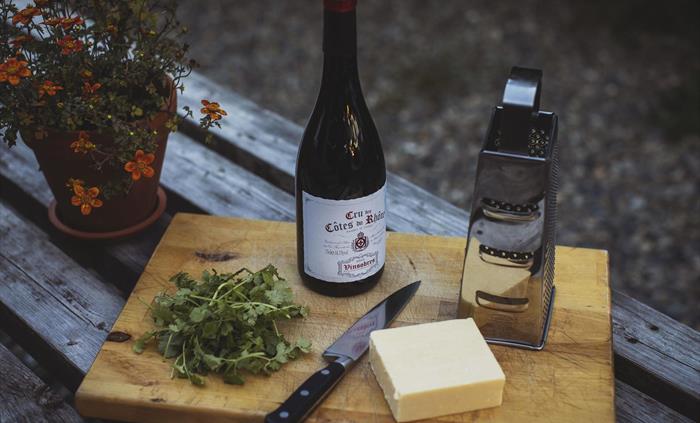 A close up of an outdoor table scene.  a wooden table with a chopping board on which there is a knife, some chopped fresh herbs, a block of cheese, a